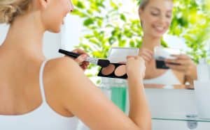 beauty, make up, cosmetics, morning and people concept - close up of happy young woman with makeup brush and contouring highlighting palette at bathroom over green natural background